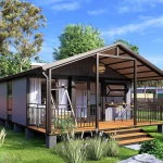 A modern expandable container home with dark metal siding, a pitched roof, and a large wooden deck, set in a sunny suburban backyard.